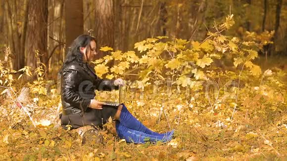 一个穿着蓝色靴子的黑发女人在阳光明媚的秋日坐在公园里拿着笔记本电脑远程工作视频的预览图