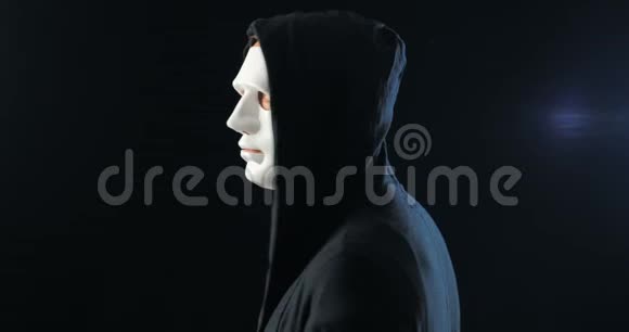 匿名者戴着白色面具遮住了他的脸一个黑色的引擎盖转过身来看着相机黑色深色背景视频的预览图