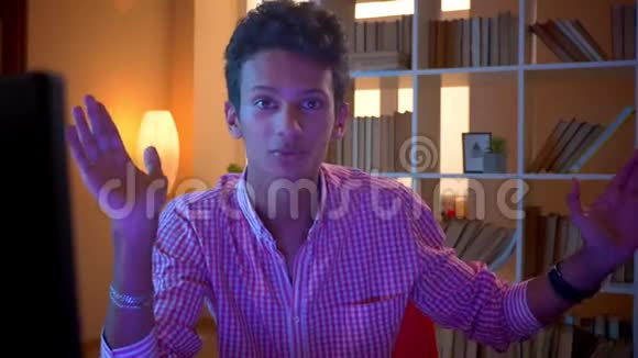 年轻迷人的印度快乐男性博客写手在一间舒适的公寓里用相机和手势进行近距离拍摄视频的预览图