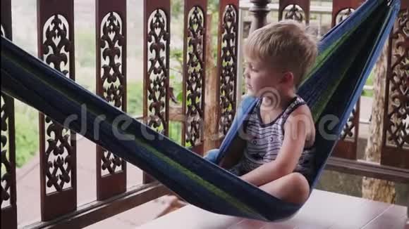 一个快乐的小男孩躺在吊床上玩耍夏天小男孩躺在阳台上的吊床上视频的预览图