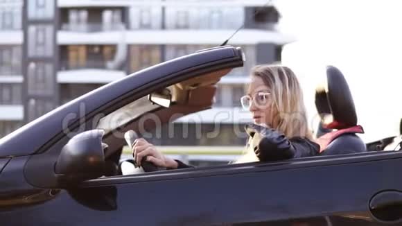 时尚年轻的金发女人驾驶她美丽的黑色运动敞篷车侧慢镜头伸出一只手视频的预览图
