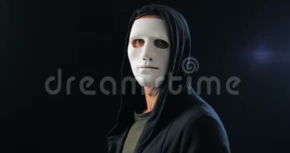 匿名者戴着白色面具遮住了他的脸一个黑色的引擎盖转过身来看着相机黑色深色背景视频的预览图