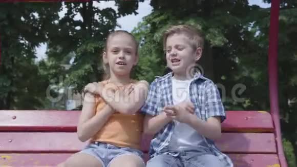 可爱的男孩和可爱的女孩坐在公园里的秋千上跳舞在户外看着相机玩得很开心A视频的预览图