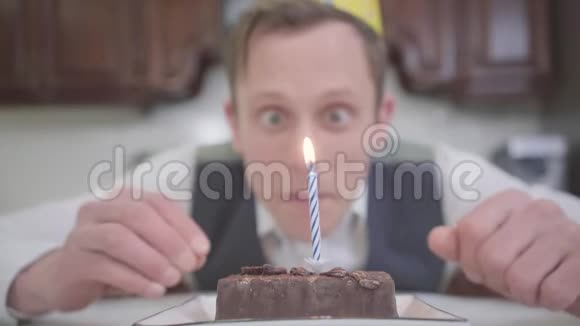 一个戴着生日帽的滑稽快乐的年轻人在厨房的一个小巧克力蛋糕上吹灭蜡烛的肖像这就是视频的预览图