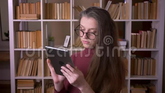 在大学图书馆里一位戴着眼镜在平板电脑上打字的白种人年轻女学生的特写照片视频的预览图