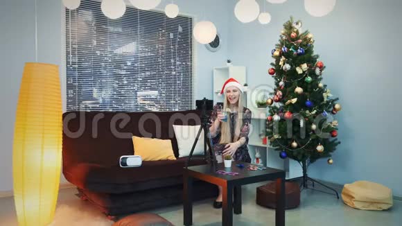 戴着圣诞帽的漂亮女孩在三脚架上用智能手机录制视频vlog视频的预览图