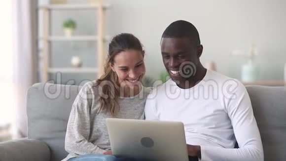 快乐的混合种族年轻夫妇使用笔记本电脑坐在沙发上视频的预览图