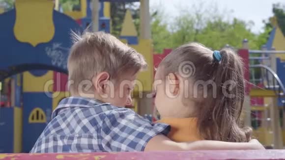 可爱的金发男孩和一个漂亮的女孩坐在操场前的长凳上拥抱一对幸福的孩子视频的预览图