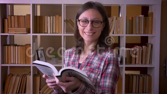 一位成功的年轻迷人的戴眼镜的女学生的特写镜头她在看一本书微笑着看着相机视频的预览图