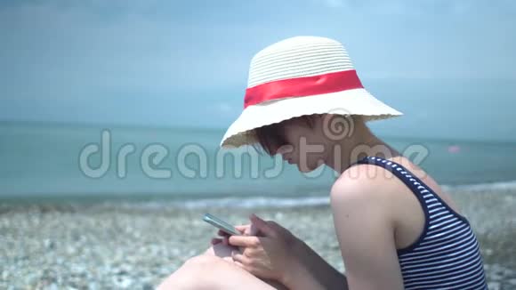 年轻的美女戴着复古的帽子穿着带蓝色和白色条纹的泳衣在孤独的夏季海滩上使用智能手机视频的预览图
