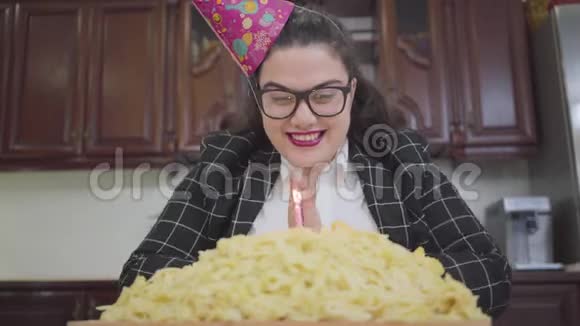 肖像可爱胖快乐的女孩戴着节日的帽子戴着时髦的眼镜坐在厨房的桌子旁吹着一个视频的预览图
