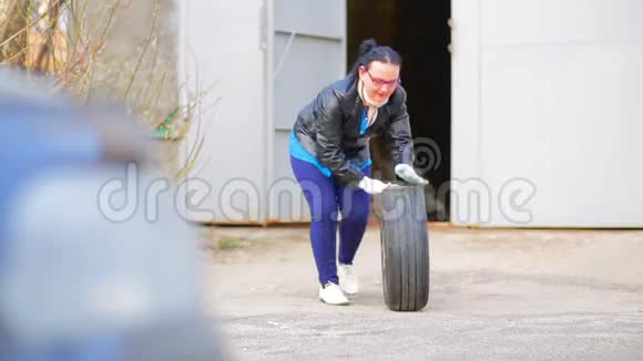 一个穿皮夹克的女人正在从车库里滚出一个带圆盘的汽车轮胎视频的预览图
