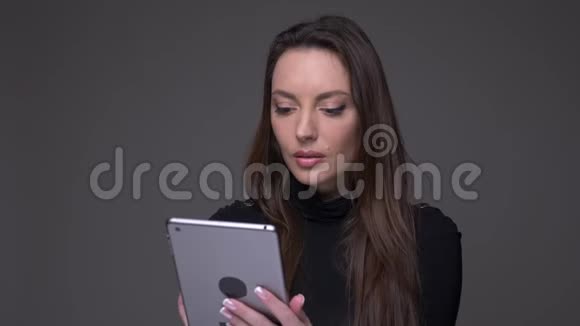 一幅美丽的黑发女人拿着平板电脑对着镜头严肃而固定地孤立在灰色上的画像视频的预览图