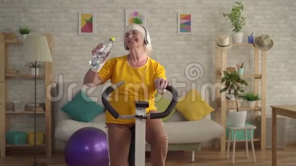 戴耳机的运动老妇人在运动自行车上进行有氧训练从瓶子里喝水视频的预览图