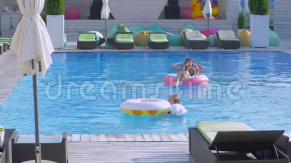 快乐的朋友在夏天的游泳池里年轻的女人在游泳池边放松女朋友带着充气环跳到水旁边视频的预览图