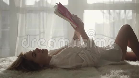年轻的白人女孩躺在床上看书的肖像穿着白衬衫的金发美女在卧室休息视频的预览图