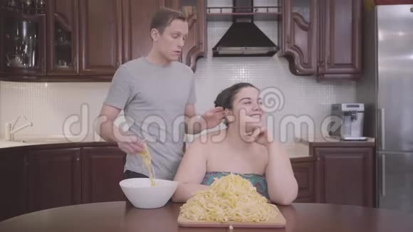 肖像可爱丰满的女人坐在家里的厨房里那个苗条的金发男人把面条挂在他肥胖的耳朵上视频的预览图