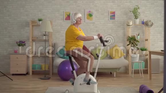 积极的祖母耳机听音乐骑着一辆静止的自行车视频的预览图