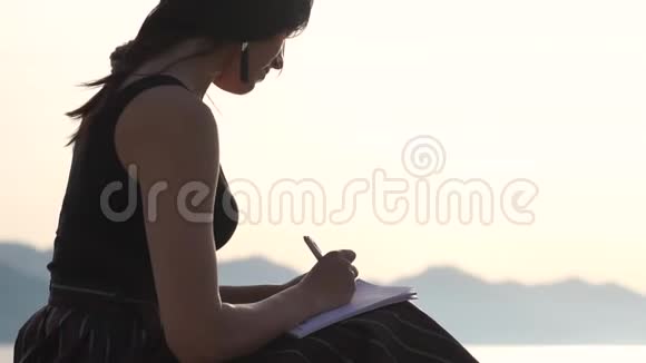 年轻女子在笔记本上写字日出时坐在湖边慢动作视频的预览图