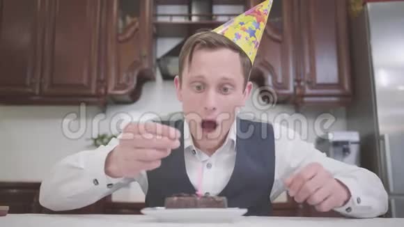 一个戴着生日帽的悲伤的年轻人模糊地描绘了厨房里一个小巧克力蛋糕上的蜡烛这就是视频的预览图
