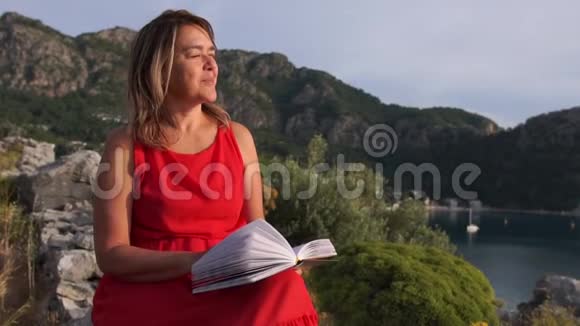 微笑的女人看着一本书坐在海面上的岩石上慢悠悠的动作视频的预览图
