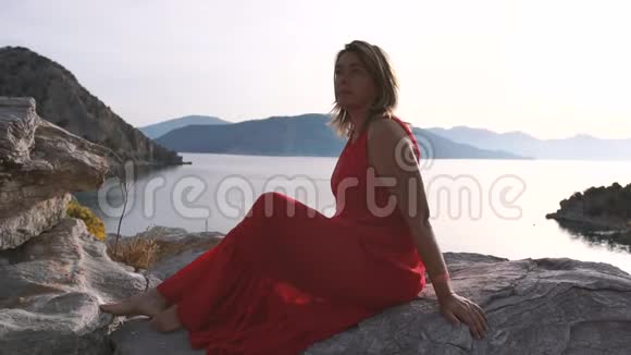 美丽的成年女人穿着红色的裙子坐在高高的海面上的岩石上视频的预览图