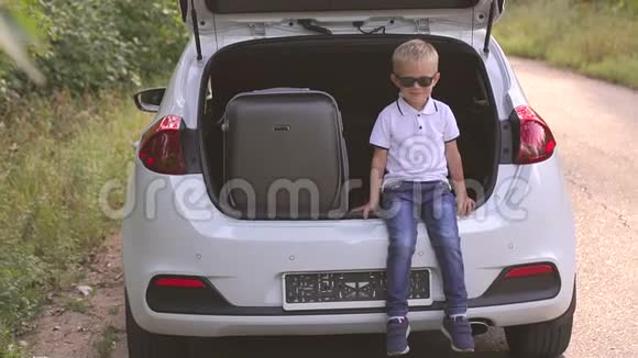一个戴太阳镜的小男孩在行李箱旁边的一辆汽车的后备箱里的肖像视频的预览图