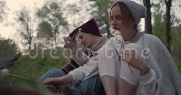 一对年轻夫妇坐在炉火旁吃棉花糖一个男人弹吉他一个女人笑视频的预览图