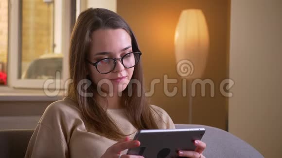 戴着眼镜的年轻白种人女性的特写肖像她用平板电脑微笑着快乐地坐在沙发上视频的预览图