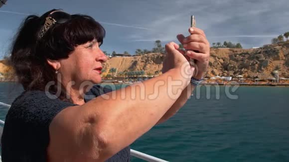 船上的老年女性快乐成年游客使用移动智能手机相机拍摄热带地区的全景照片视频的预览图