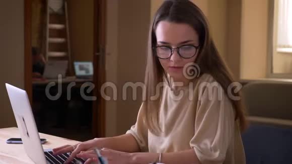 一位年轻貌美的高加索女学生戴着眼镜在大学室内的笔记本电脑上打字视频的预览图