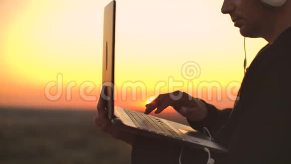 在日落时分拿着笔记本电脑和啤酒站在屋顶上一个穿着连帽衫的男人在享受乐趣和沉思视频的预览图