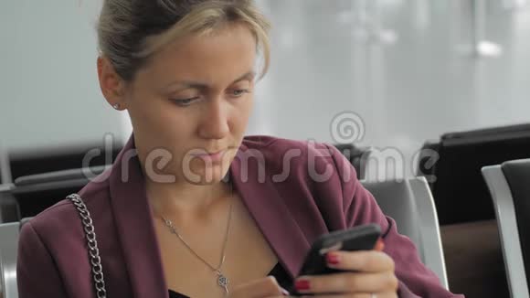 漂亮的白种人女性在智能手机上打字而坐在机场航站楼深思熟虑和专注视频的预览图
