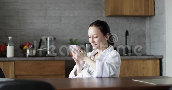 一个穿着白色浴袍的成年女人坐在厨房里手里拿着手机视频的预览图