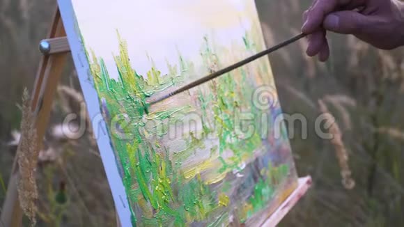 男性艺术家在自然景观中的空地上描绘日落景观画布和画笔动作的近景视频的预览图