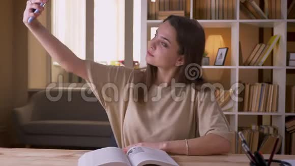 一位年轻迷人的白种人女学生在室内阅读一本书并在电话中自拍的特写照片视频的预览图