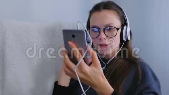 戴着眼镜和耳机的年轻女子正在智能手机上听音乐用卡拉OK唱一首歌视频的预览图