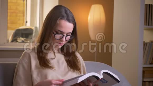 一位年轻迷人的女学生在舒适的公寓里坐在沙发上看书的特写镜头视频的预览图