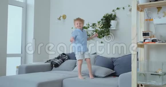 一个4岁的男孩留着白色的头发和一件蓝色的衬衫在沙发上跳跃和微笑在家玩得很开心普兰克和视频的预览图
