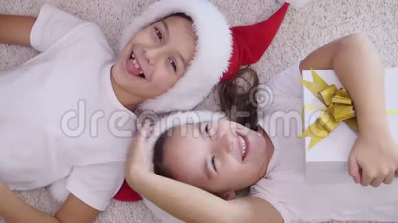两个孩子穿着圣诞帽和睡衣头对头躺在地毯上肚子上放着圣诞礼物盒视频的预览图