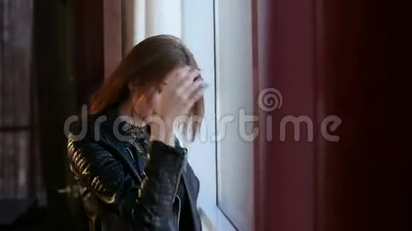 一个穿着皮夹克的美丽悲伤的女孩站在窗户附近视频的预览图