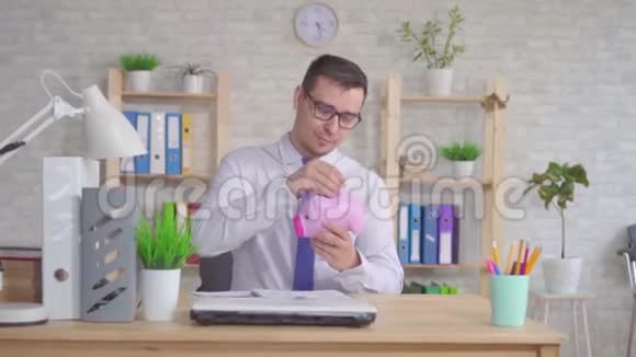 男人坐在办公室里把一枚硬币扔进储蓄罐看着相机伸出手指视频的预览图