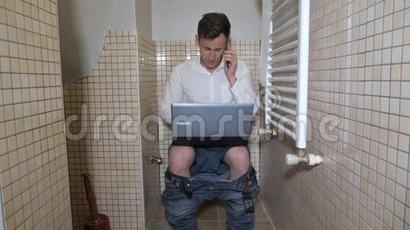 年轻人工作狂正在厕所里工作用电脑在智能手机上聊天视频的预览图