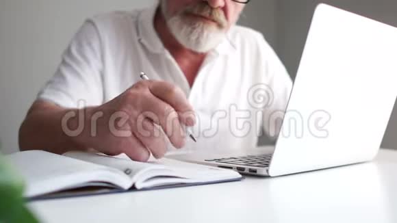 留着灰色胡子的商人在办公室用笔记本电脑工作用铅笔在笔记本上做笔记远程视频的预览图
