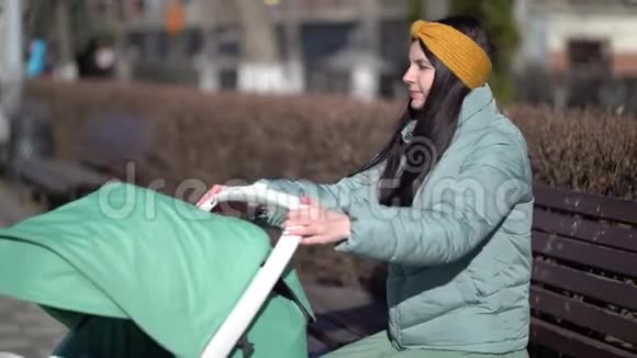 年轻母亲带着新生儿户外她坐在长凳上婴儿睡在婴儿车里视频的预览图