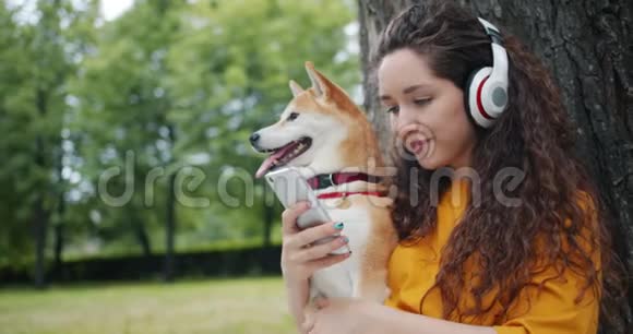 戴着智能手机耳机的快乐女士然后在公园拥抱宠物狗视频的预览图