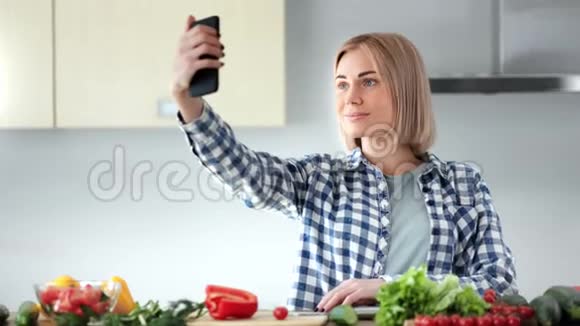 美丽的年轻家庭主妇在烹饪健康食品时使用智能手机自拍视频的预览图