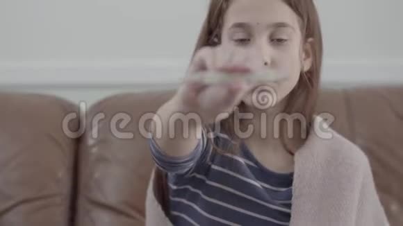 描绘年轻疲惫的生病女孩坐在沙发上对着镜头展示一个温度计生病孩子的概念医药视频的预览图