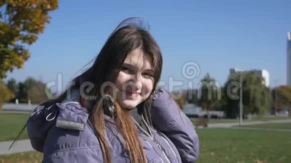 公园里的女孩穿着羽绒服的迷人女孩走在秋天的公园里微笑着抚摸着她的头发视频的预览图