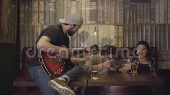 年轻的大胡子男子在酒吧弹吉他他的朋友坐在附近喝啤酒在酒吧休闲伙计们视频的预览图